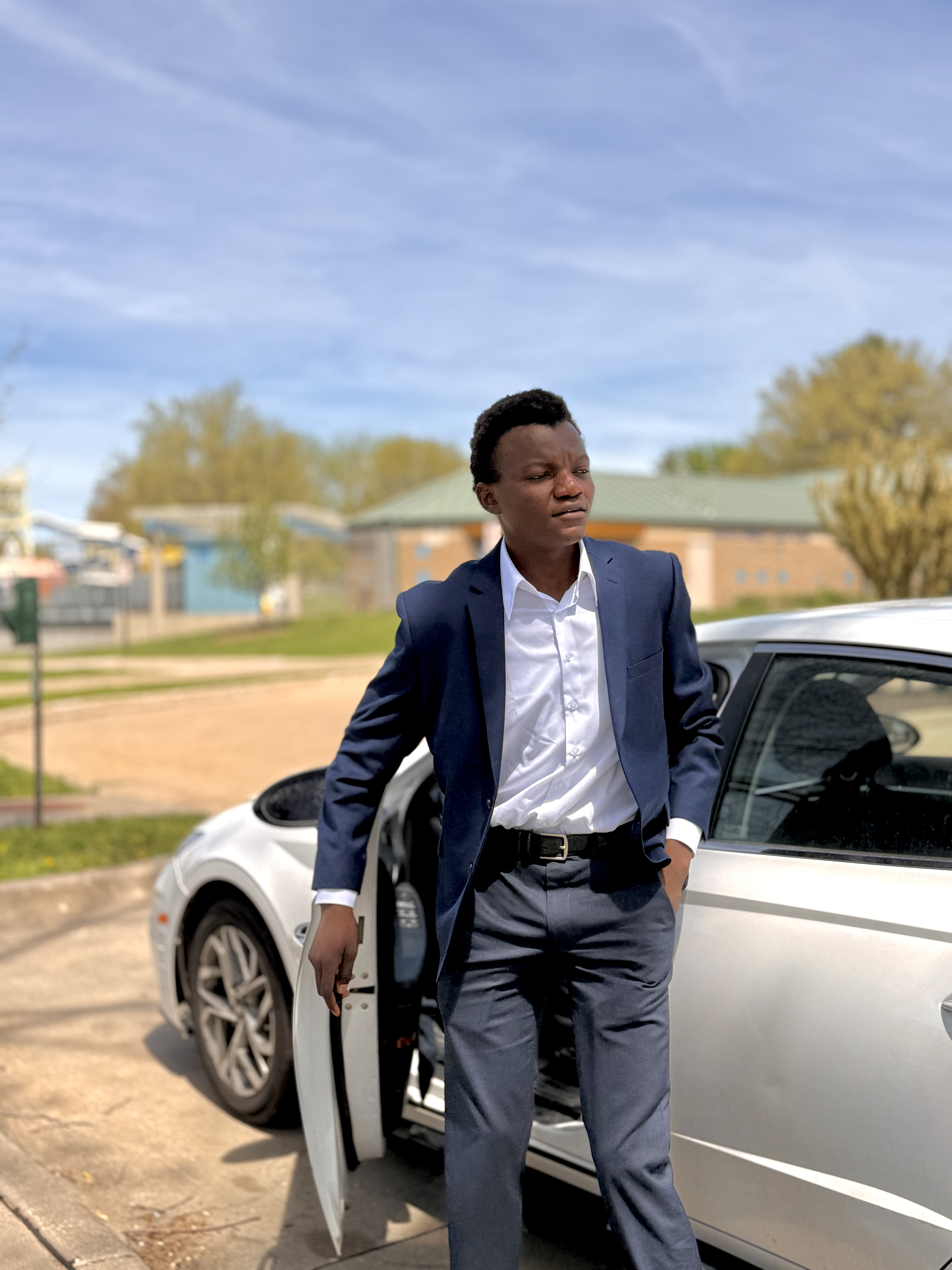 Jeune homme noir en costume bleu marine debout à côté d'une voiture blanche ouverte.